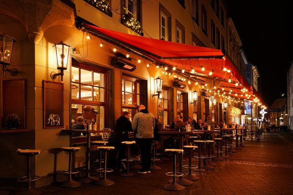 Kornelia Brinkman-Rodden - Annahme Beleuchtetes Restaurant mit Terrasse und roten Markisen in einer nächtlichen Stadt.