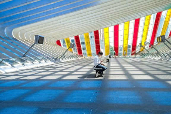 Liege 2 - Annahme - Helmut Oberritter Person sitzt in einer modernen Halle mit bunten Streifen im Dach.