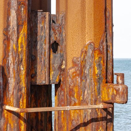 Rost 01 Rostige Metallstruktur an einem Wasserstand mit der Küste im Hintergrund.