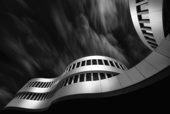 Juli 2025 - Ute-Christa Scherhag Welleförmiges modernes Gebäude mit geschwungener Fassade und dramatischem Himmel.