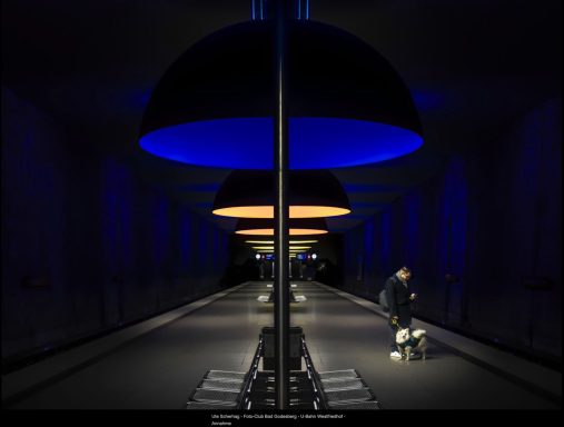 LaFo 2025 - Annahme - Ute Scherhag Dunkler Bahnhof mit blauen und orangefarbenen Leuchten, eine Person sitzt auf einem Stuhl.