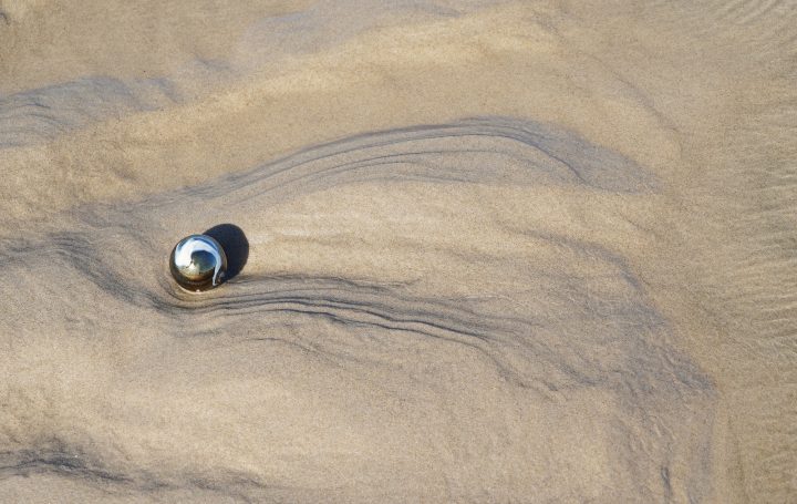 Sand -2- Eine einzelne Kugel liegt auf sandigem Untergrund, umgeben von sanften Wellen.