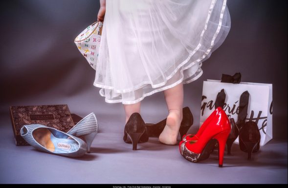 Ute Scherhag - Annahme Eine Person trägt schwarze Schuhe, umgeben von roten High Heels und verschiedenen Taschen.