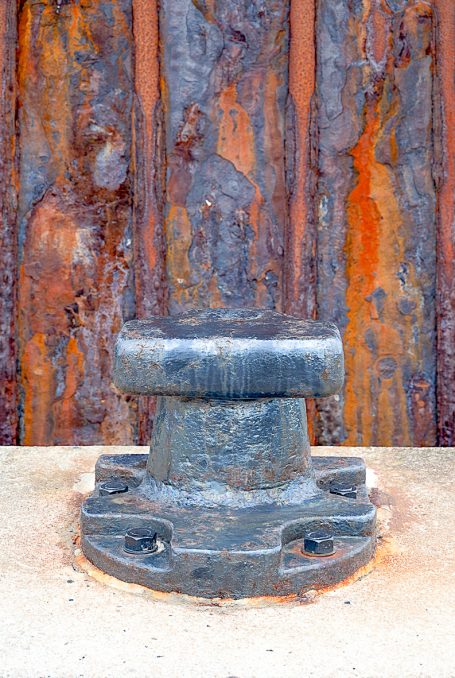 Rost 03 Ein rostanhaftes Bolzen mit einer runden Basis vor einer rustikalen Wand.