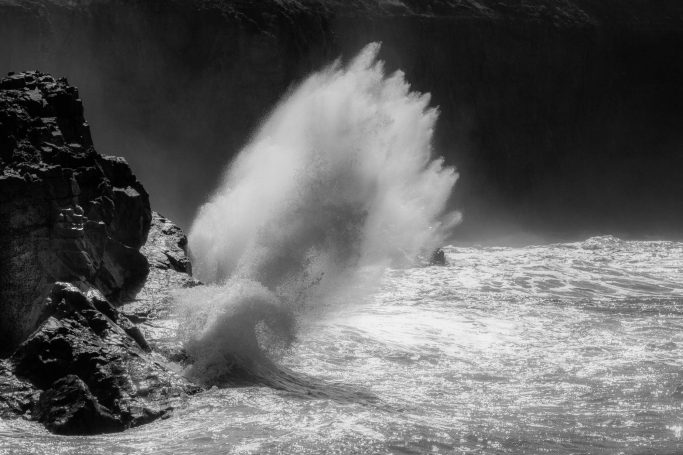 Mai 2025 - Uwe Müller Spritzendes Wasser trifft auf Felsen am Ufer, monochrome Darstellung.