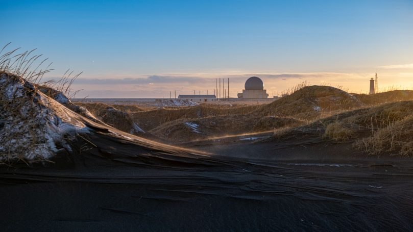 Hügelige Sanddünen im Vordergrund mit einem Gebäude und einem Kuppelturm im Hintergrund.