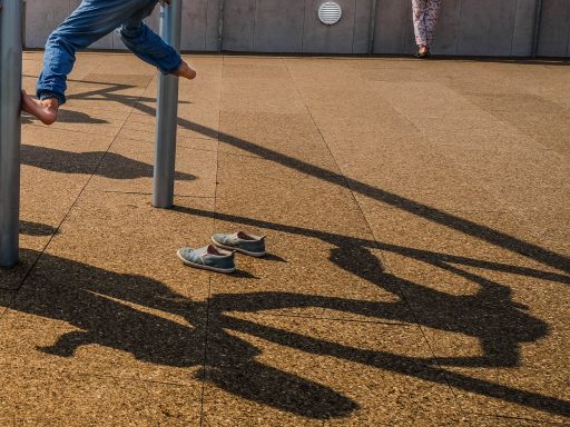 Schatten - Annahme - Helmut Oberritter Ein Kind schwingt an einem Spielgerät, Schuhe liegen auf dem Boden, Schatten sind sichtbar.